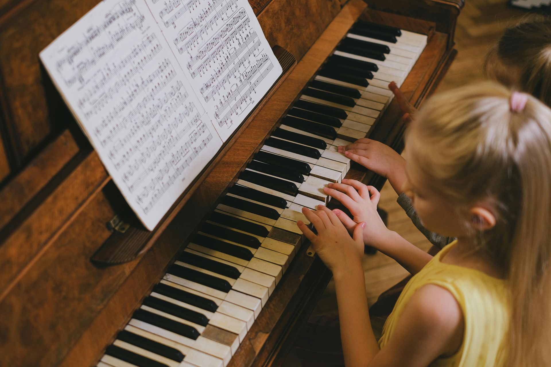 Young girl with blonde hair playing a piano, reading sheet music. Inside, brown piano, yellow shirt.