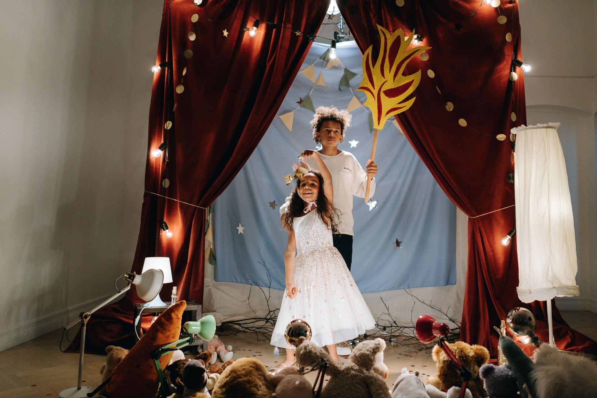 Two children on stage: girl in white dress, boy with prop. Red curtains, toy audience.