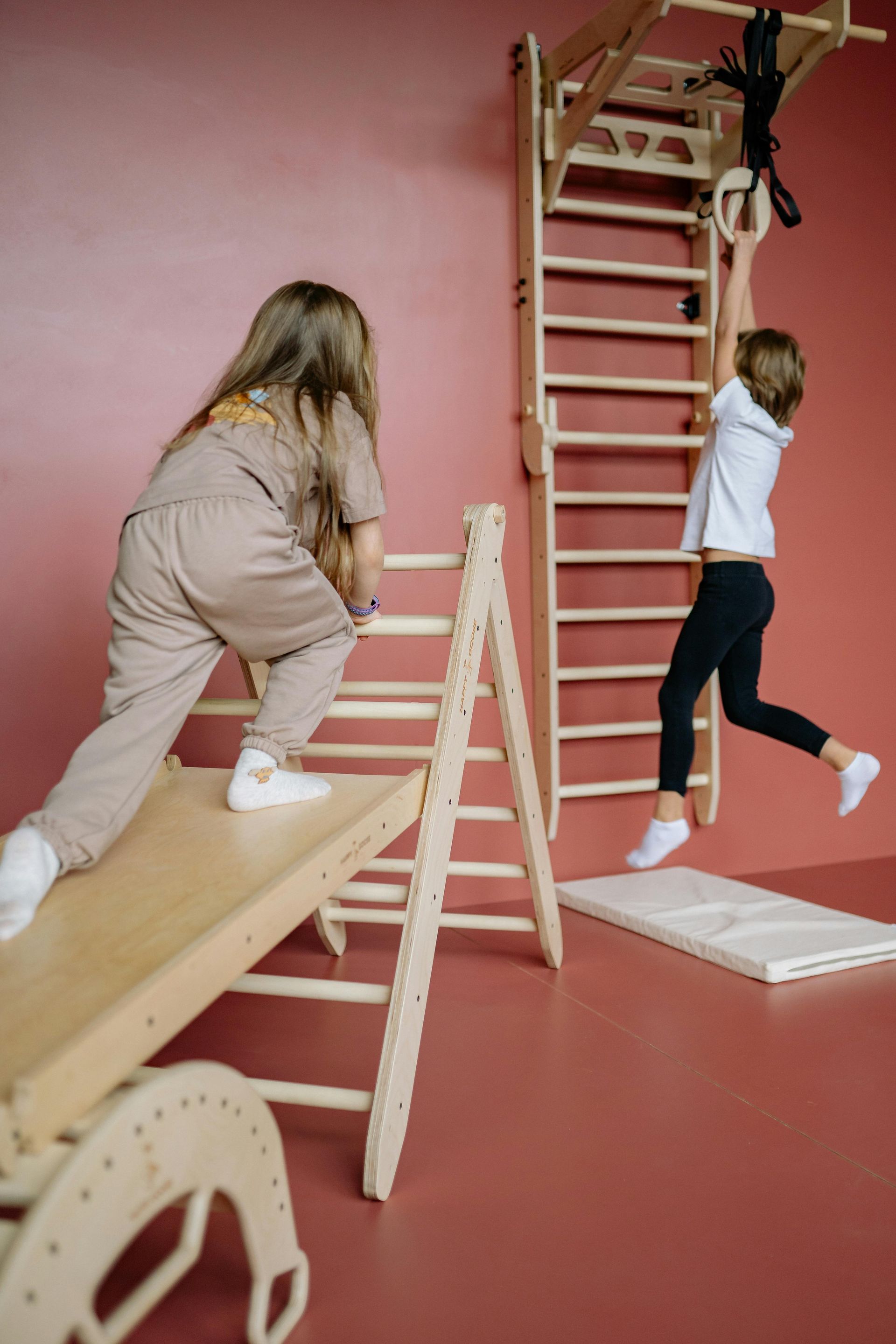 Two young girls playing on wooden climbing equipment in a red room.
