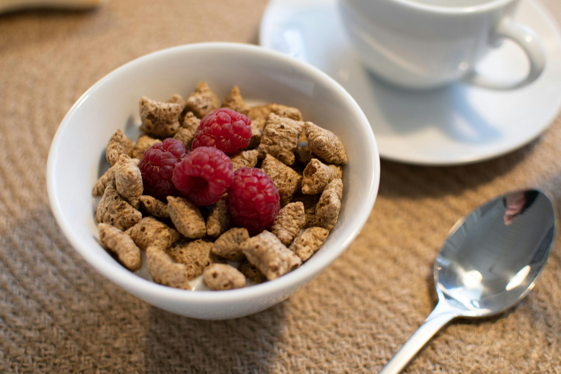 A bowl of cereal with raspberries and a spoon next to a cup of coffee.