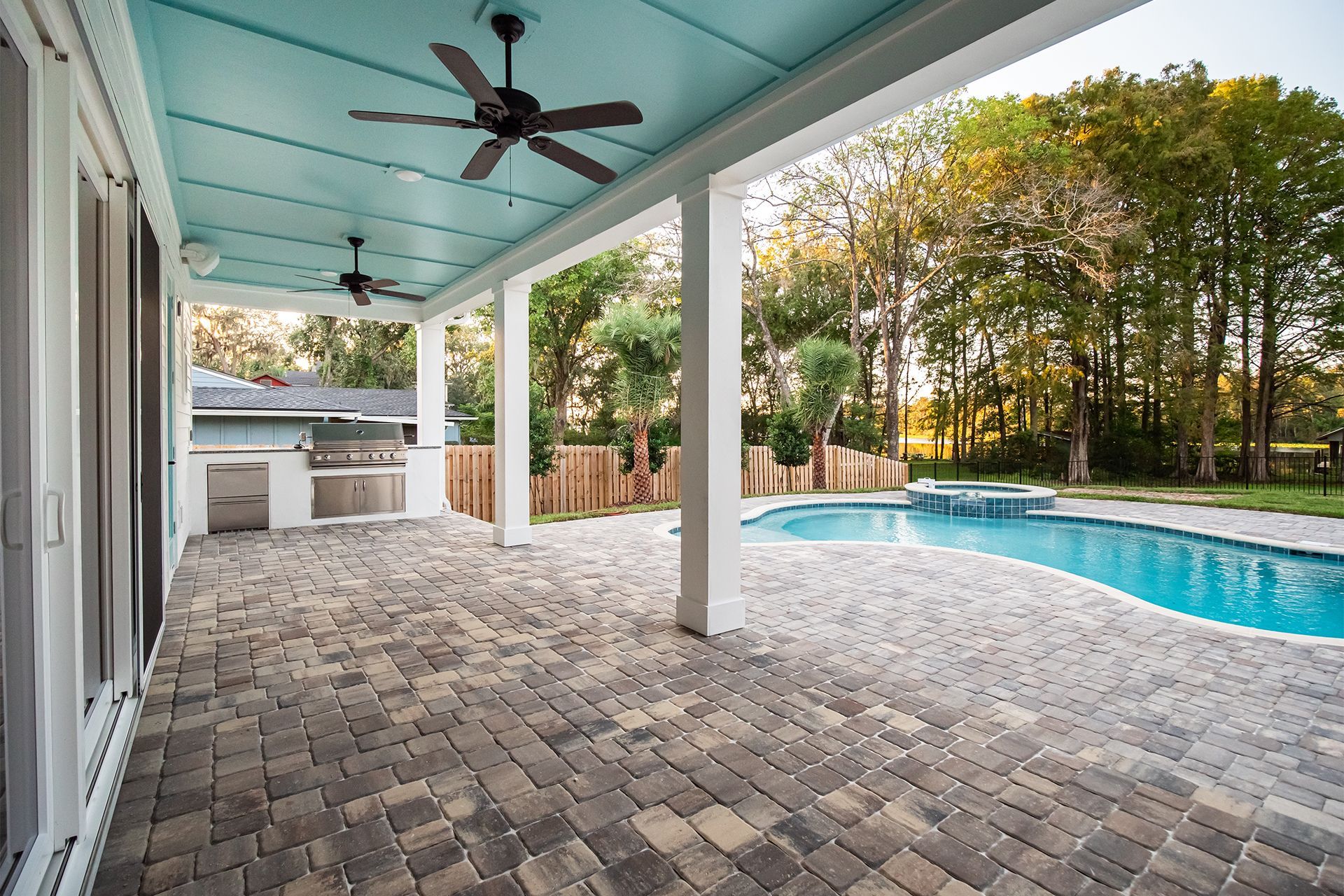 Covered patio with blue ceiling, brick pavers, outdoor kitchen, pool in background.