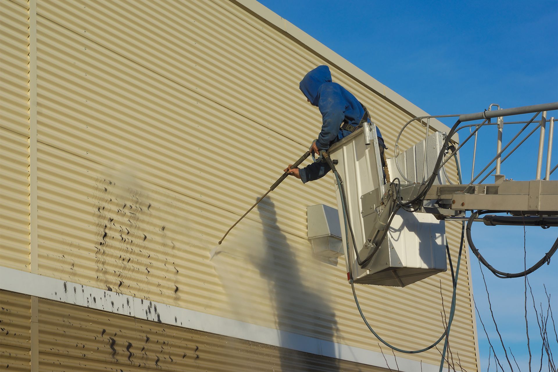 Person pressure washing a beige building exterior from a lift against a blue sky.