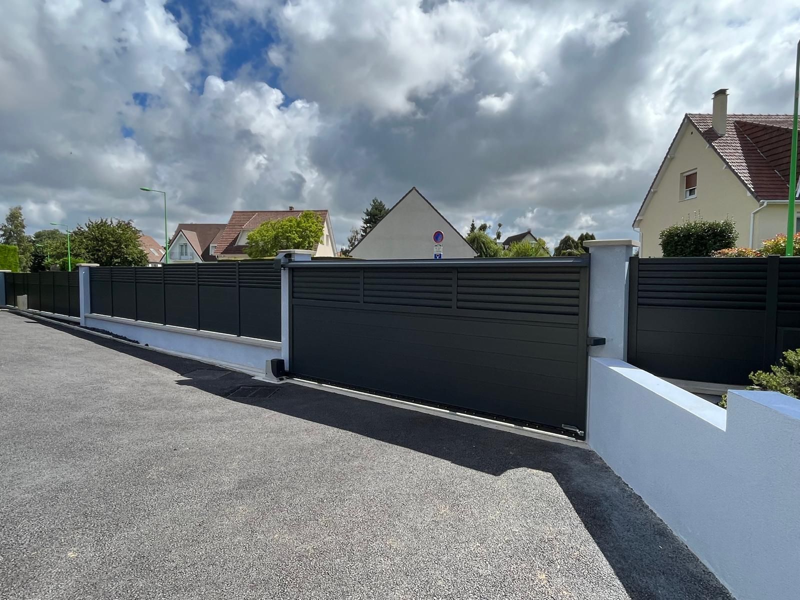 Portail coulissant noir et clôture le long d'une allée, avec des maisons et un ciel nuageux en arrière-plan.
