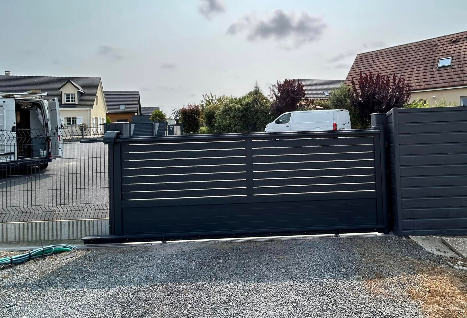 Un portail coulissant à lattes horizontales gris foncé devant une maison avec du gravier et des maisons en arrière-plan.