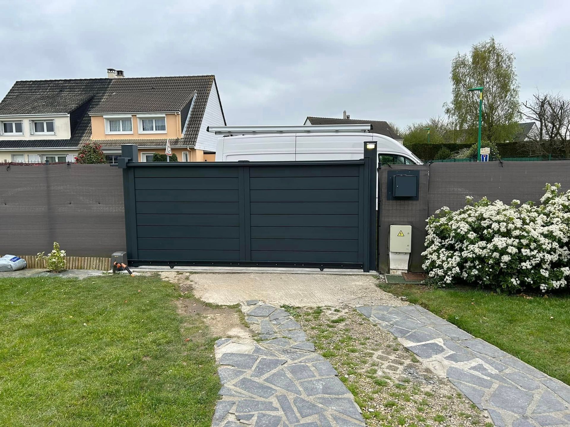 Portail double gris foncé avec une allée en pierre, menant à une propriété résidentielle derrière une clôture marron.