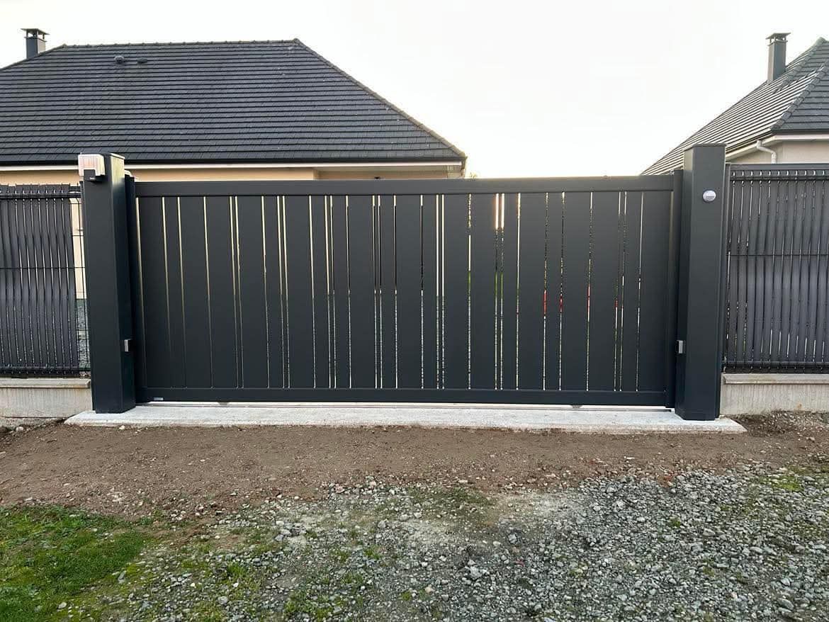 Portail d'allée moderne noir avec lattes verticales en bois, adossé à une maison au toit gris.
