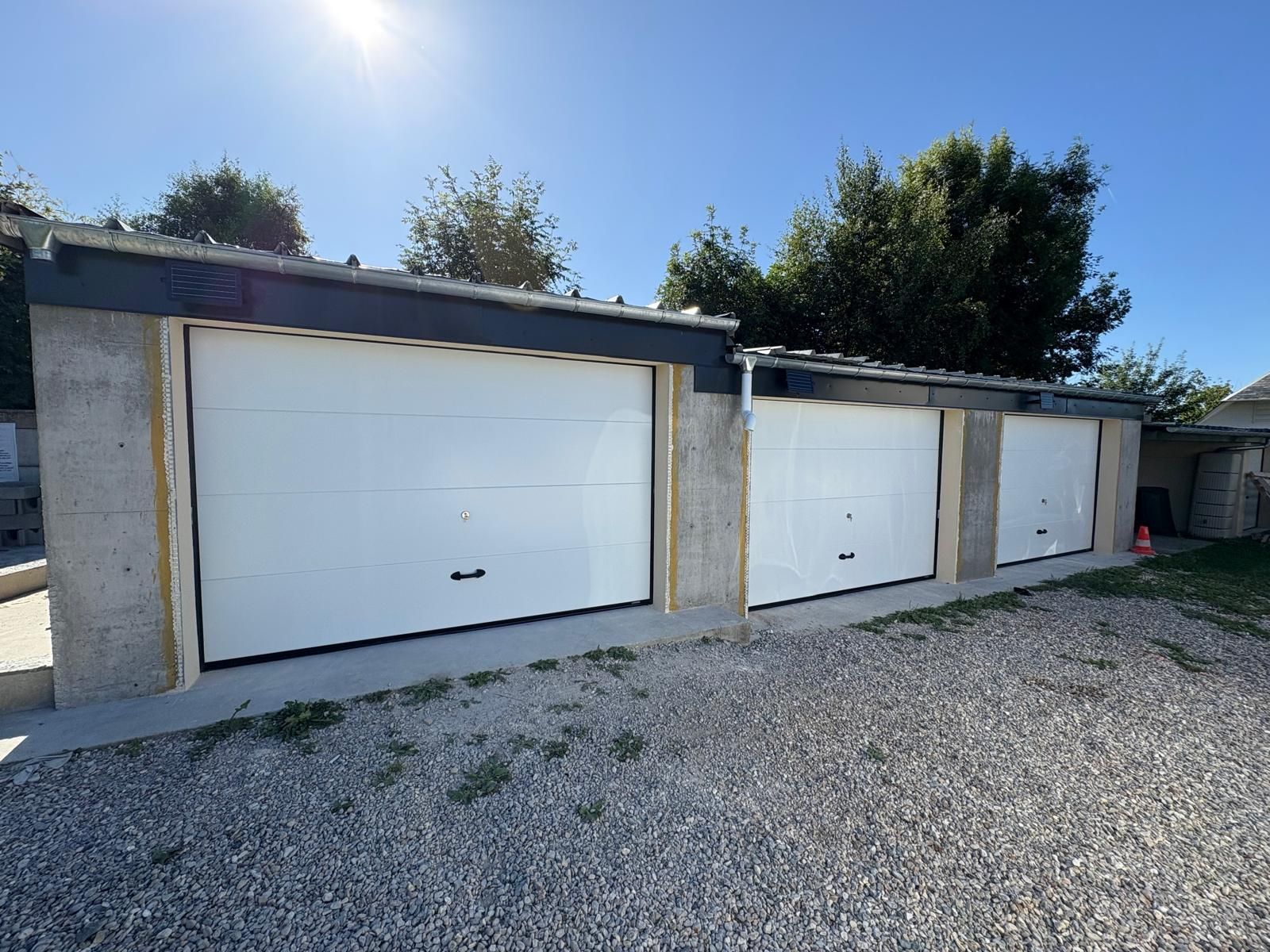 Trois portes de garage blanches alignées, avec des murs en béton et une allée en gravier, sous un ciel bleu vif.