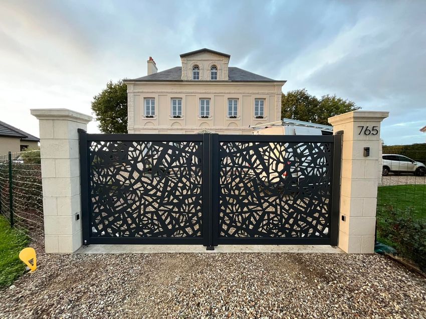 Des portes décoratives en métal noir s'ouvrent sur une allée de gravier, avec une maison en pierre claire en arrière-plan.