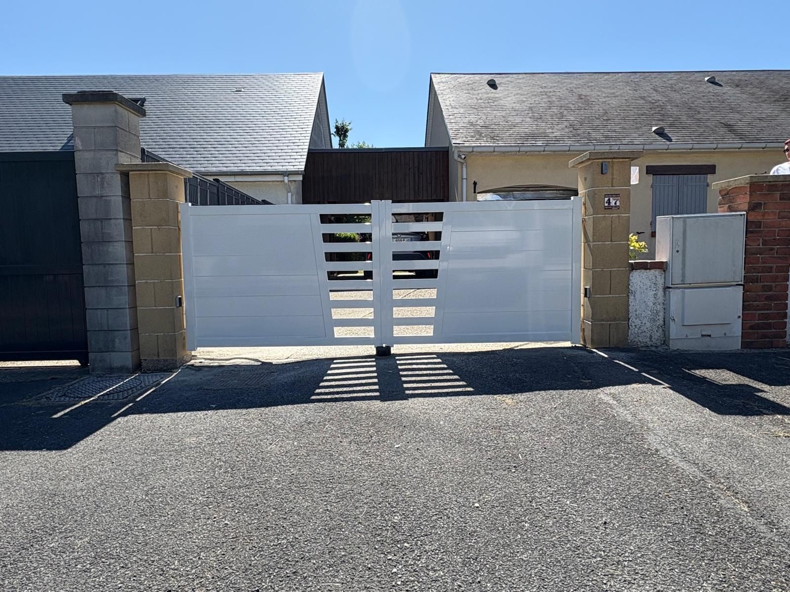 Portail coulissant blanc entre deux piliers en pierre à l'entrée d'une maison, sur fond de ciel bleu.