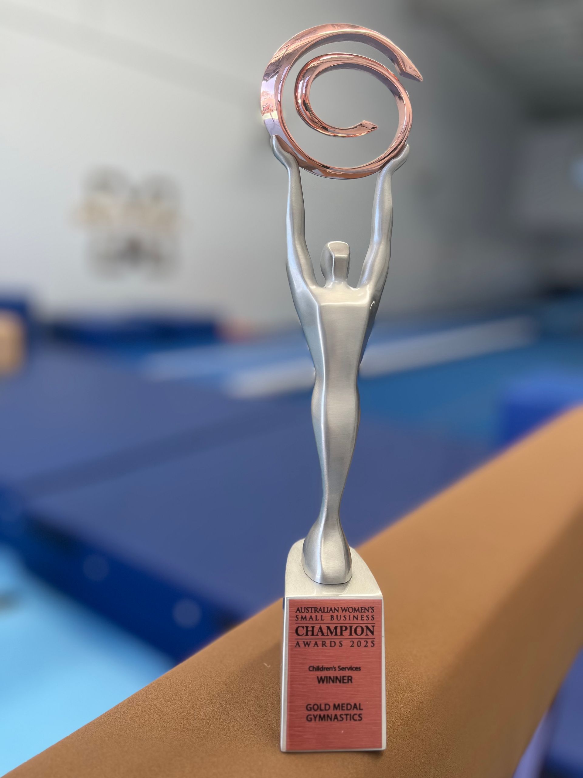 Trophy of a silver figure holding a copper spiral. It's on a pink base, set in a gymnastics gym.