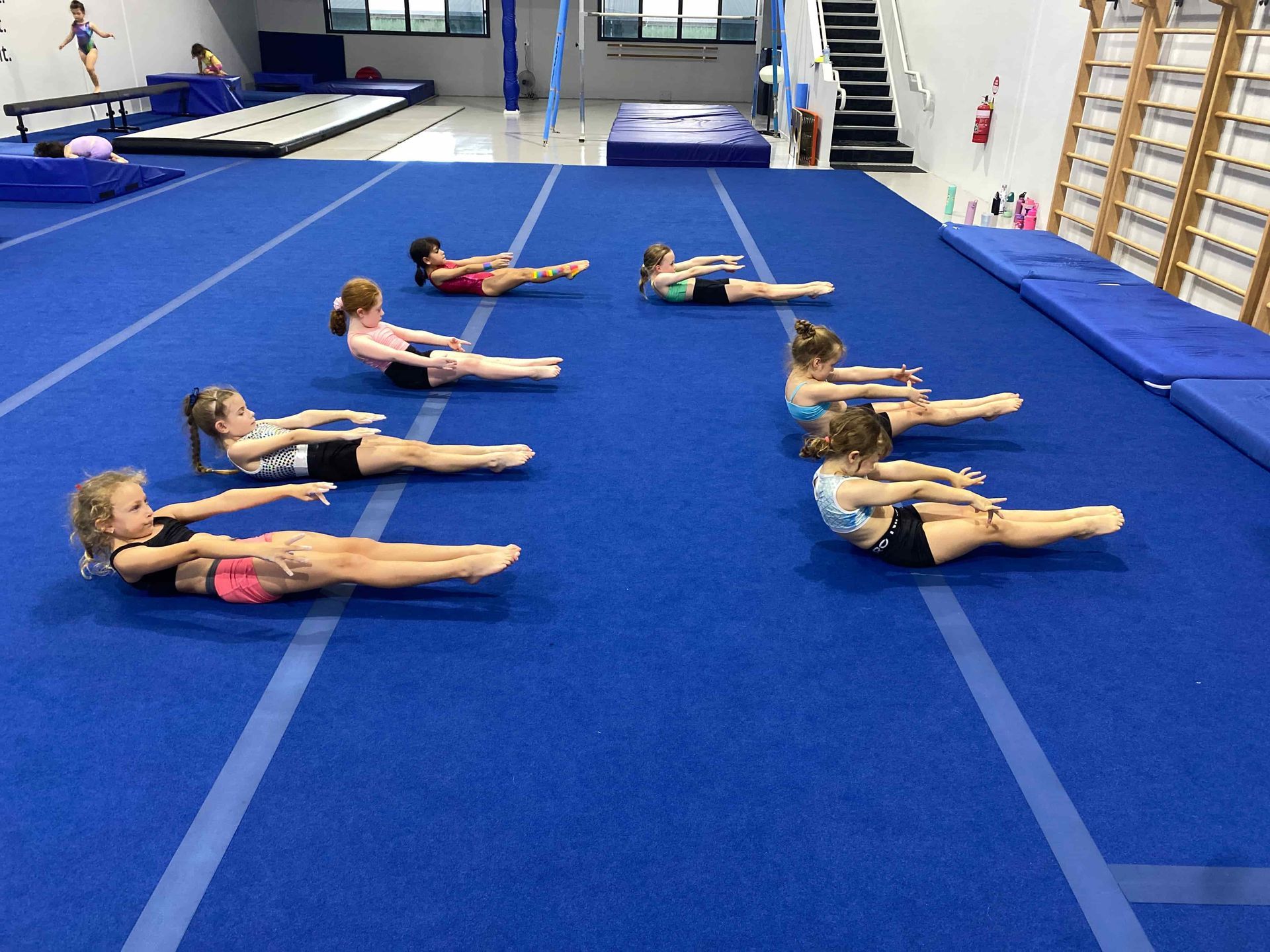 A group of young girls are doing exercises on a blue mat in a gym. Gym — Gold Medal Gymnastics In Gold Coast, QLD