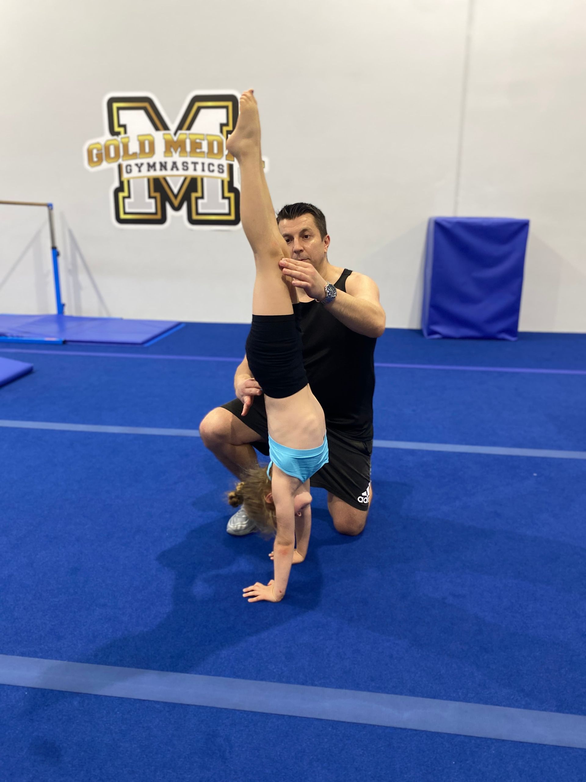 A man is helping a little girl do a handstand on a blue mat. — Gold Medal Gymnastics In Gold Coast, QLD
