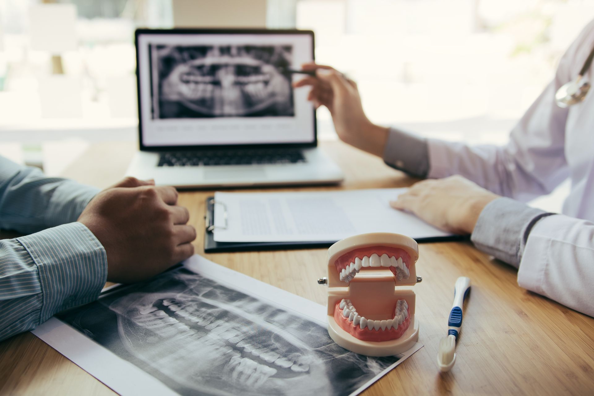 Dentist showing patient dental x-ray on laptop; model teeth, documents, and pen on desk.