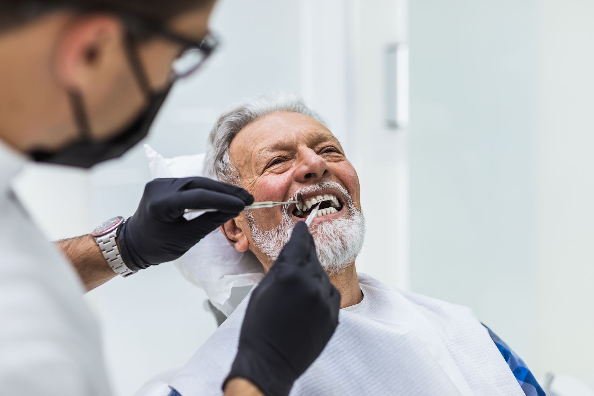 Dentist examining older man's teeth with clear tray in a bright office. Man smiles; both wear masks.