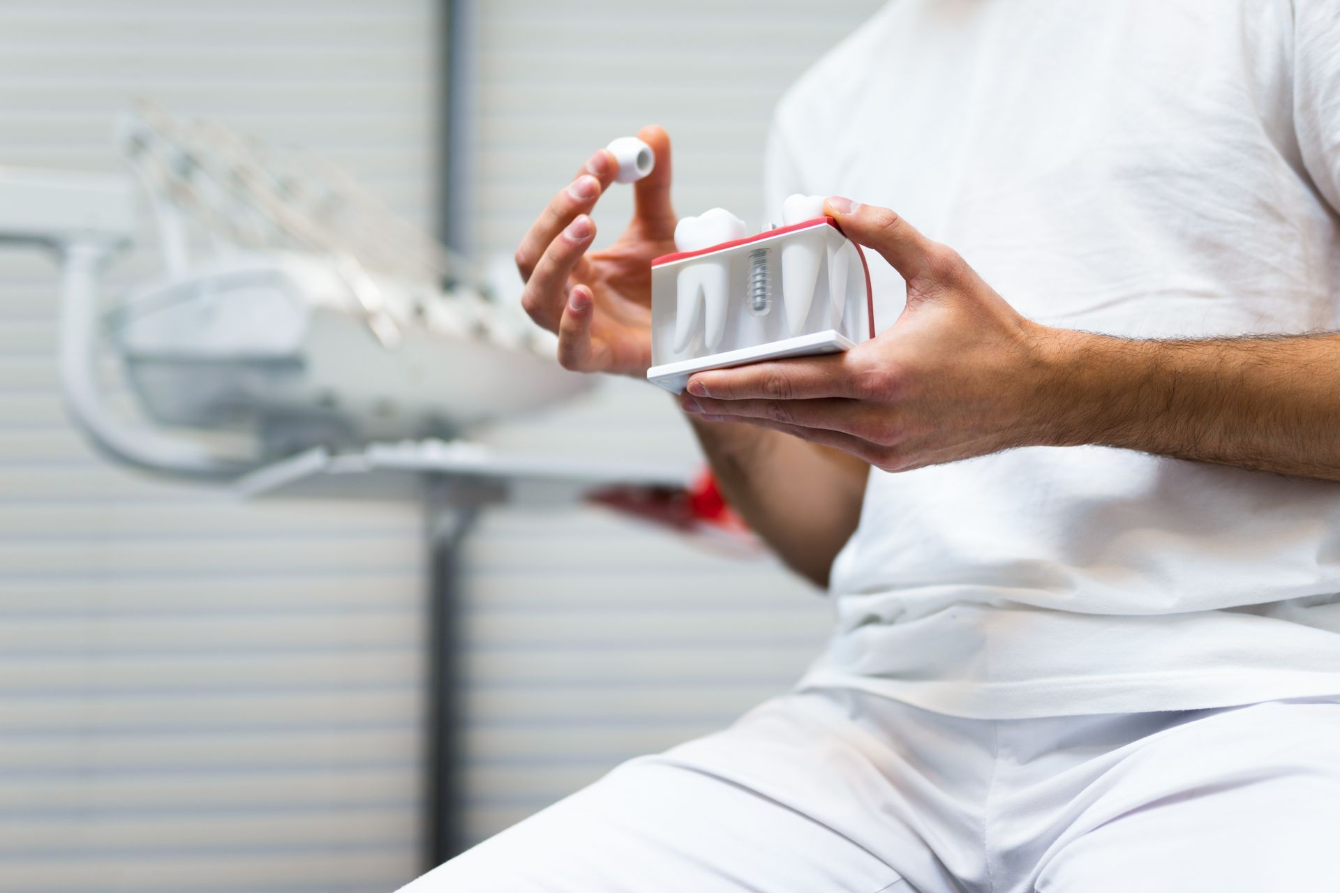 Dentist holding a small container with a small white dental filling, a dental chair in the background.
