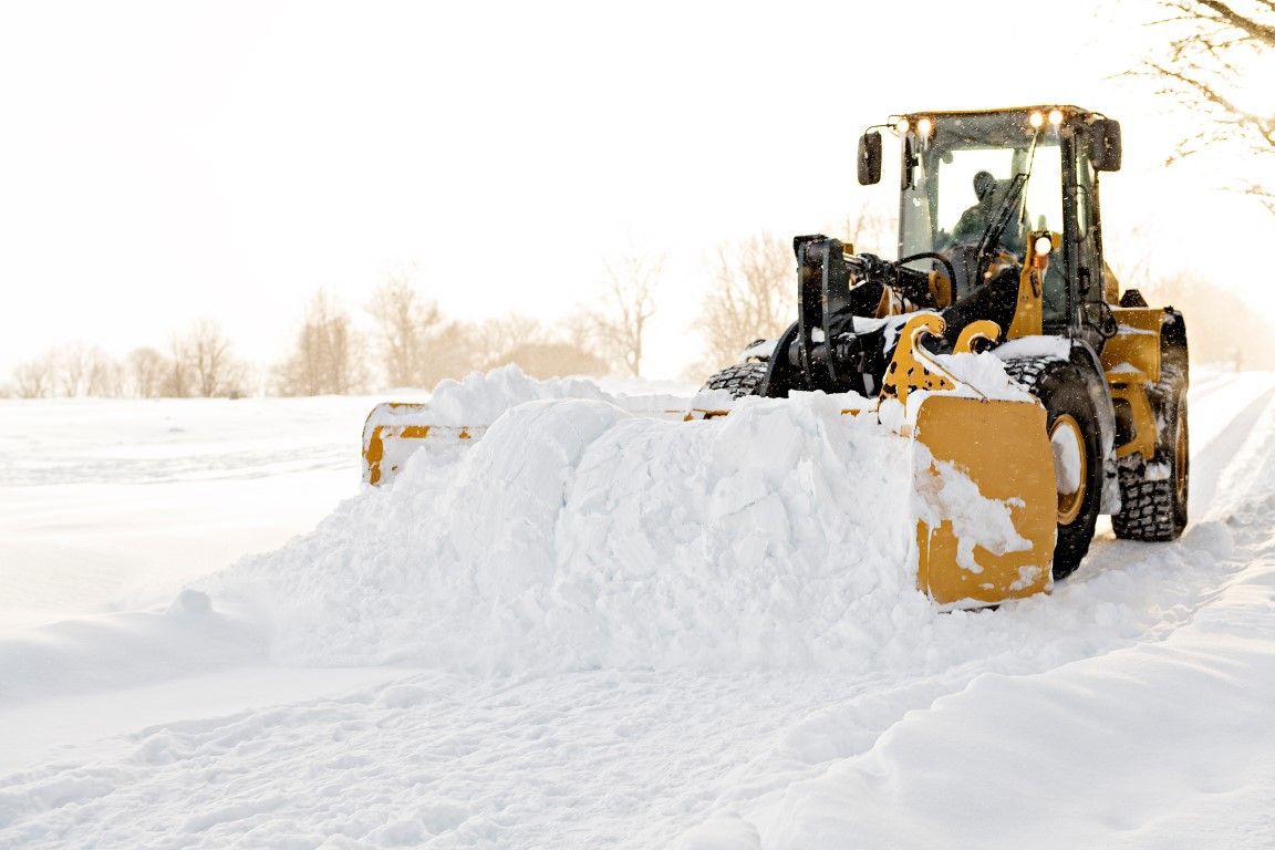 Yellow snowplow clearing snow from a road on a bright, snowy day.
