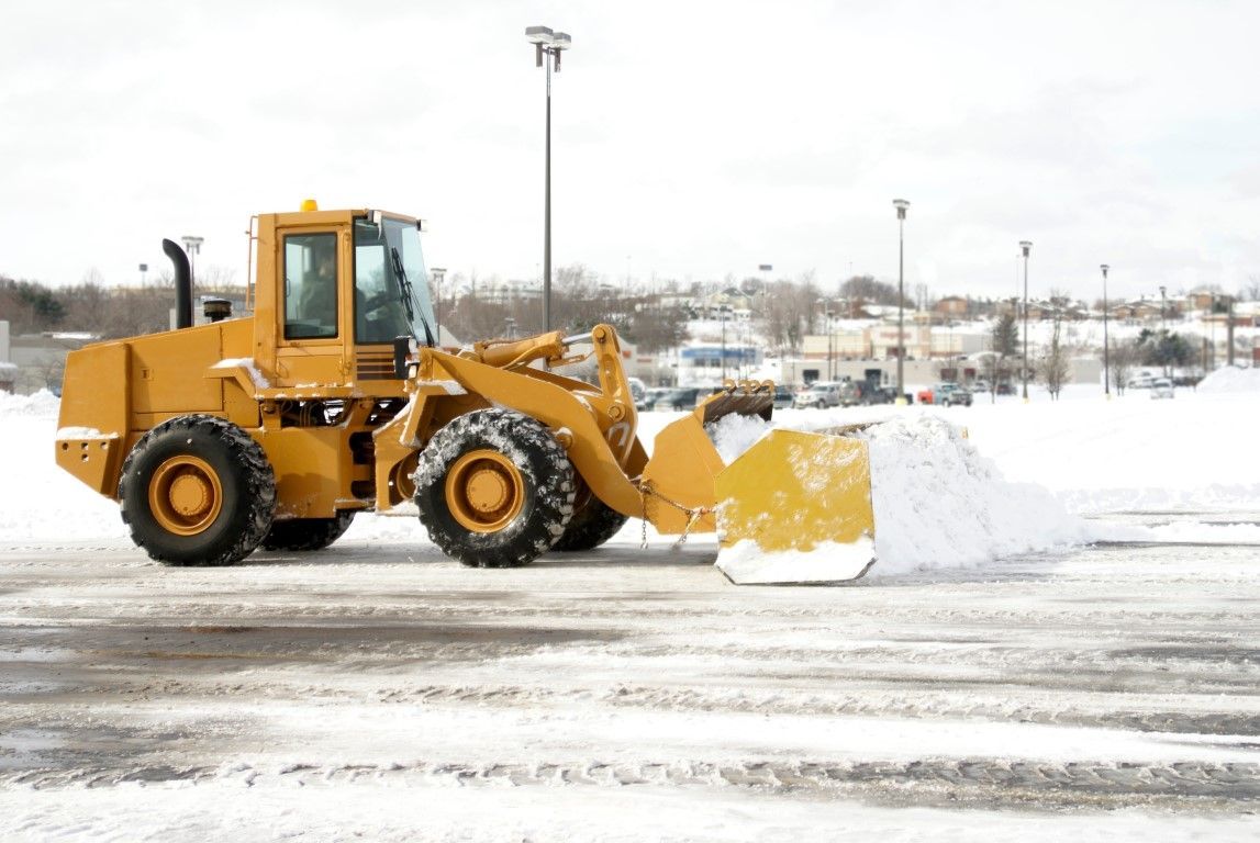 Yellow snowplow clearing snow from a parking lot on an overcast day.