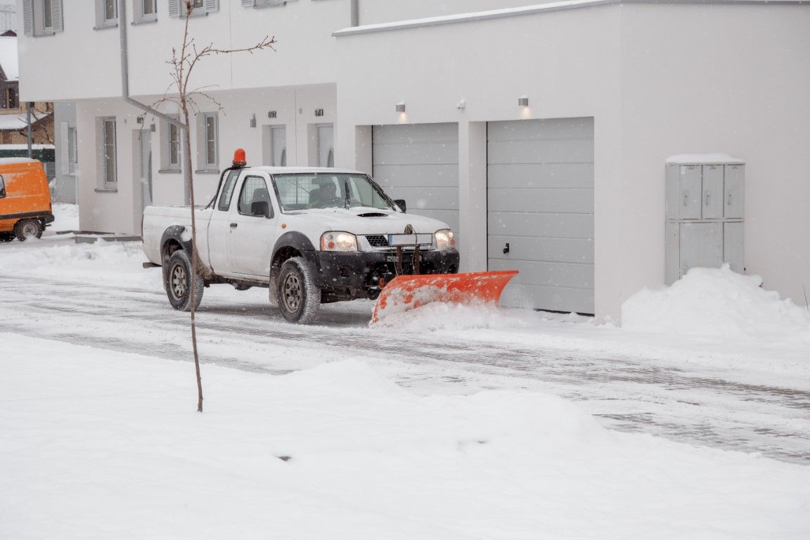 White pickup truck plowing snow from a driveway, next to a white building with garage doors.