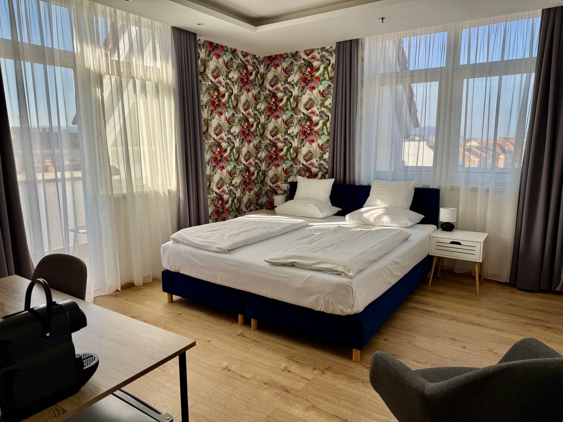 Hotel room with a bed, desk, chair, and large windows with curtains; natural light.