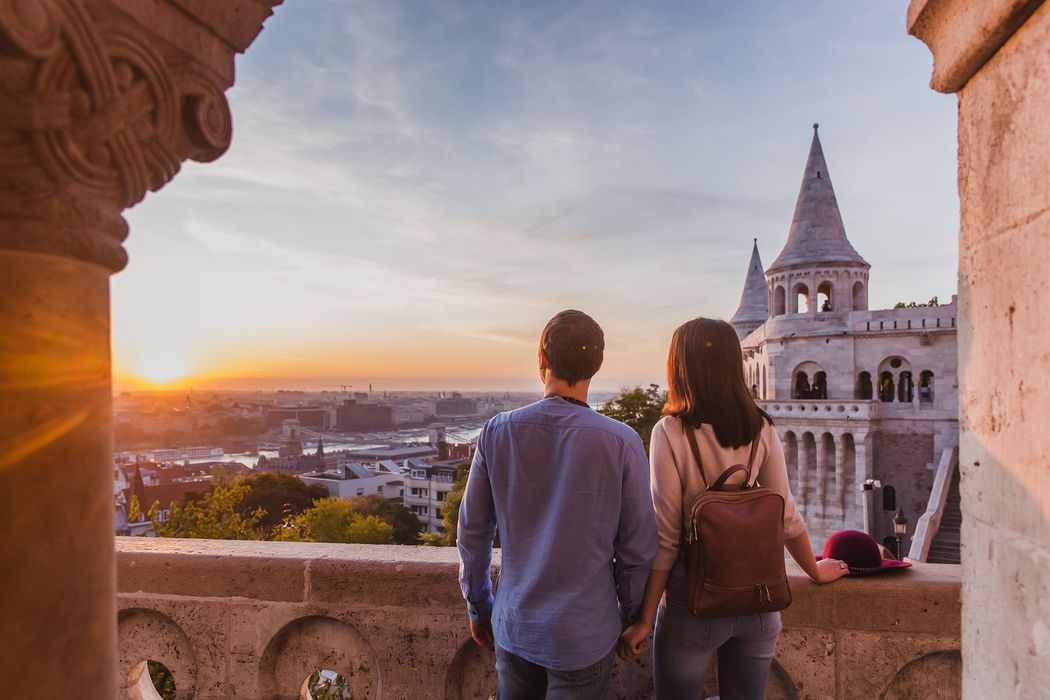 A man and a woman are standing on a balcony looking at the sunset over a city.