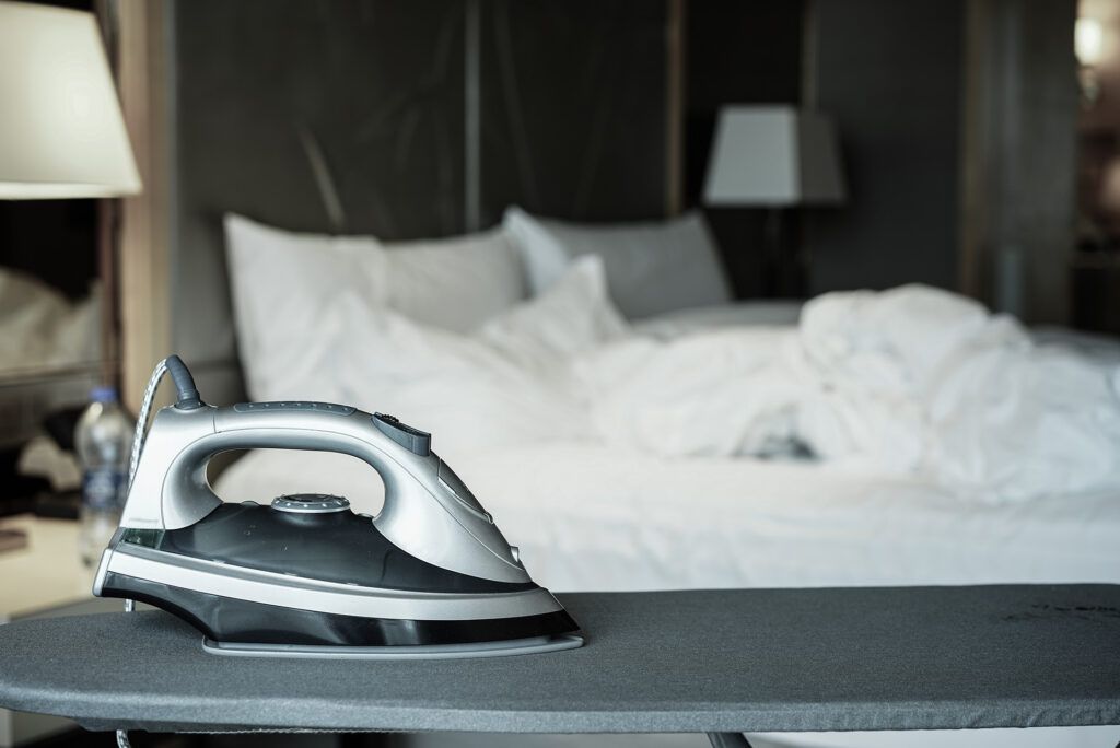 Iron on an ironing board in a hotel room with rumpled white bedding and a lamp.