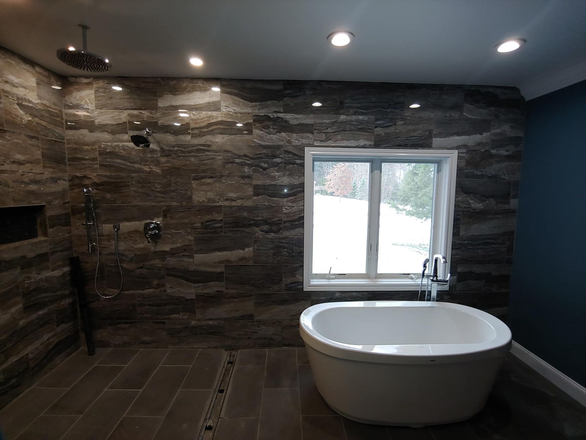 Modern bathroom with stone tile walls, rainfall shower, soaking tub by a window.