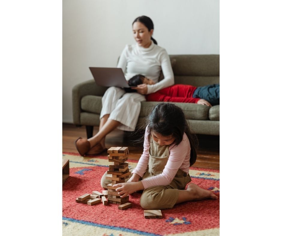 A woman is sitting on a couch while a girl plays with wooden blocks on the floor.