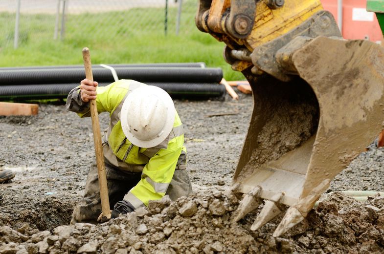 Construction worker digging with a shovel near an excavator, in muddy ground.