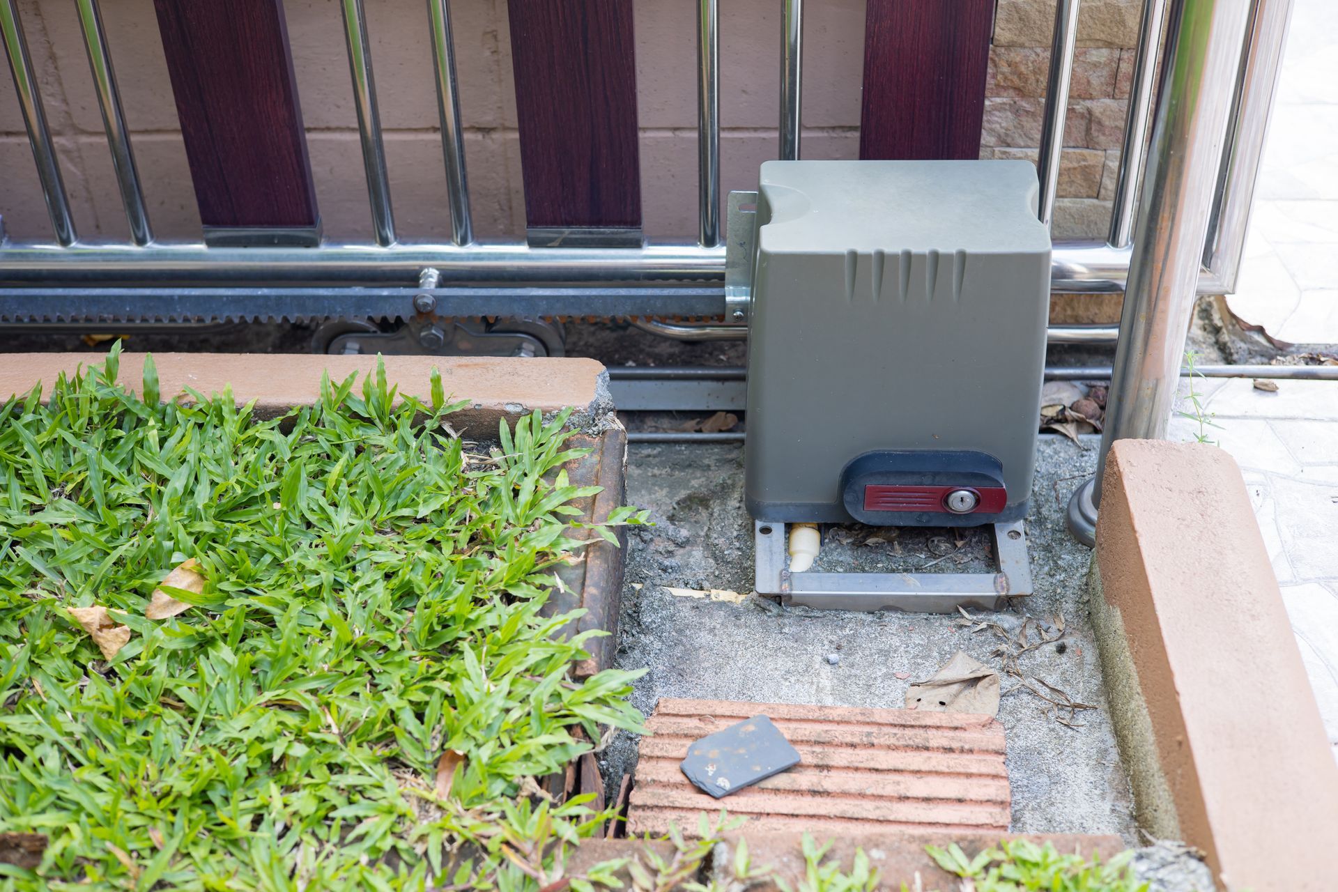 Sliding gate opener mechanism, with control panel, next to grass and brick pathway.