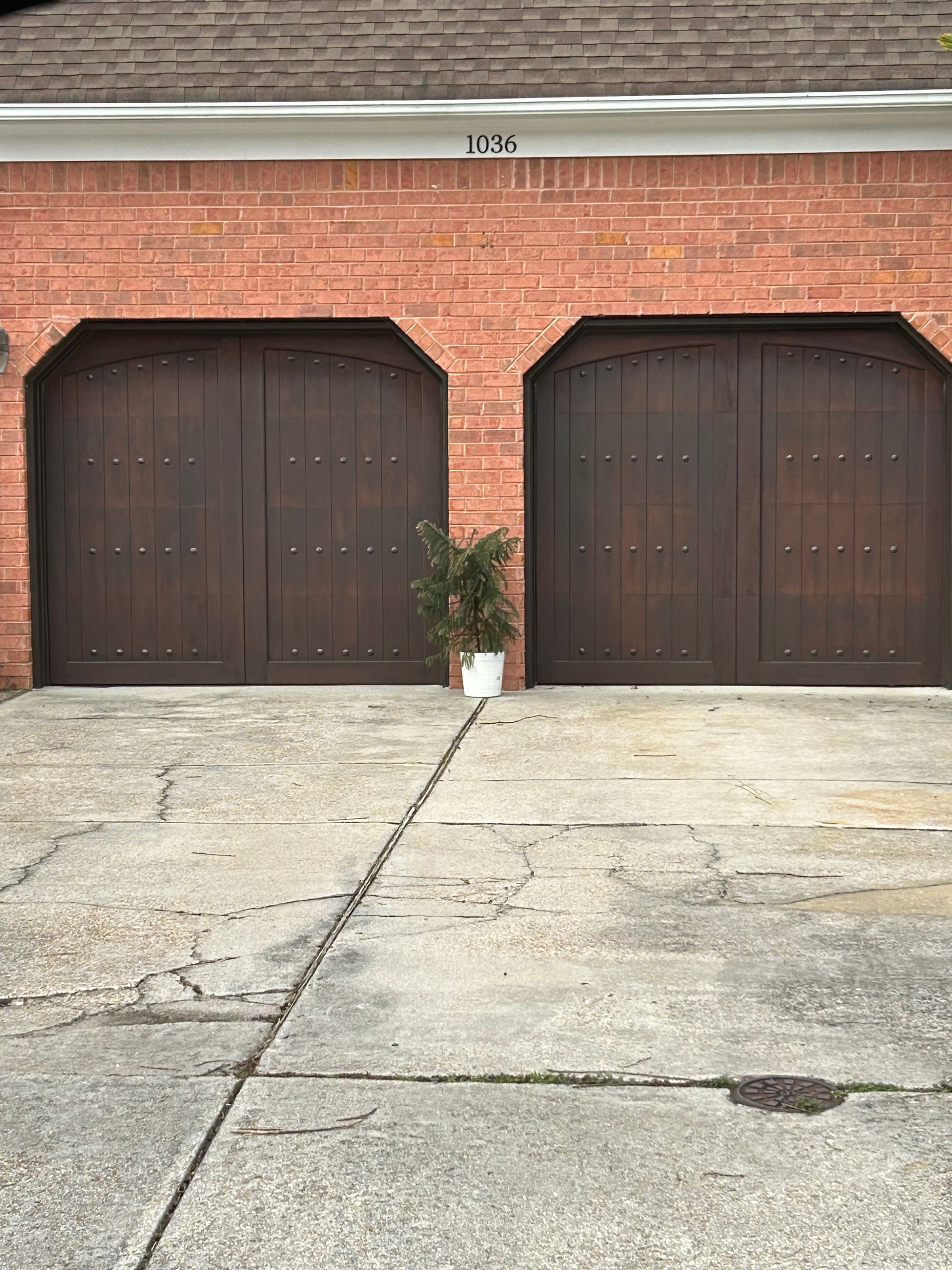 Two dark brown garage doors with brick facade, a small plant in front, and cracked concrete.