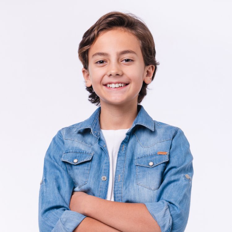 A young boy in a denim shirt is smiling with his arms crossed.