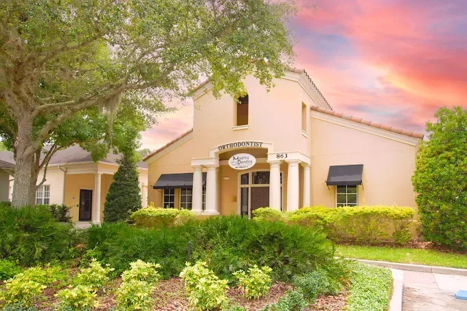 Beige stucco building with black awnings, surrounded by greenery under a colorful sunset.