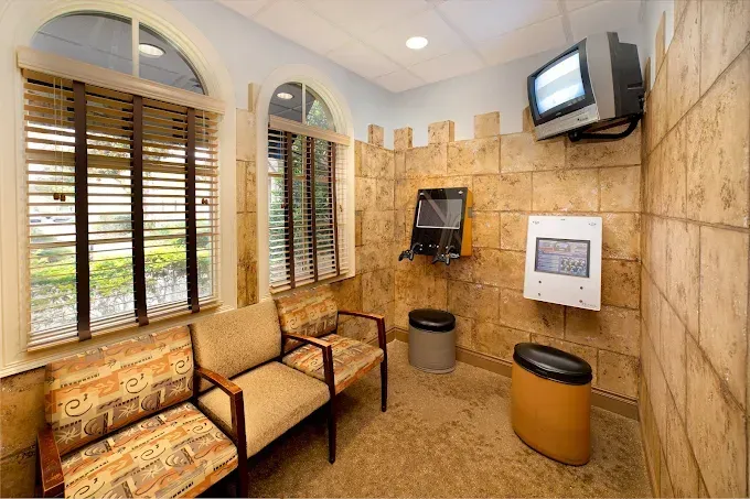 Waiting room with stone-like walls, arched windows with blinds, a mounted TV, and seating.