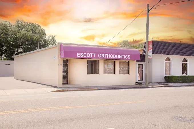Exterior view of Escott Orthodontics, a light-colored building with a purple awning against an orange and yellow sky.