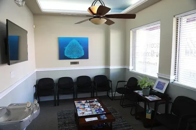 Waiting room with dark chairs, art, windows, and ceiling fan. A sink is on the left.
