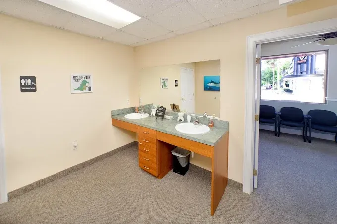 Public restroom with two sinks, mirror, and access to a waiting room. Light beige walls, neutral floor.