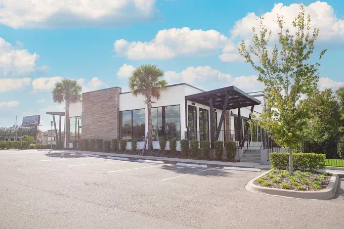Modern building with large windows, landscaping, and a parking lot against a blue sky with clouds.