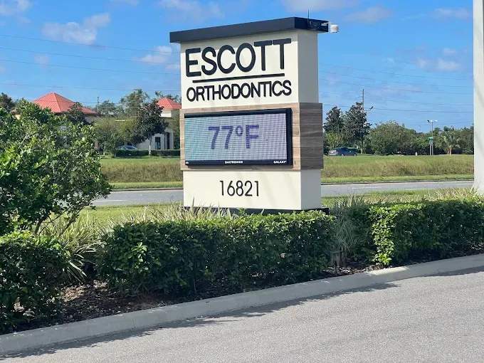 Sign for Escott Orthodontics displaying the temperature as 77°F. The sign is beige, black, and tan and sits by a green bush.
