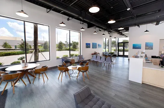 Interior of a modern waiting area with tables, chairs, large windows, and art on the walls.