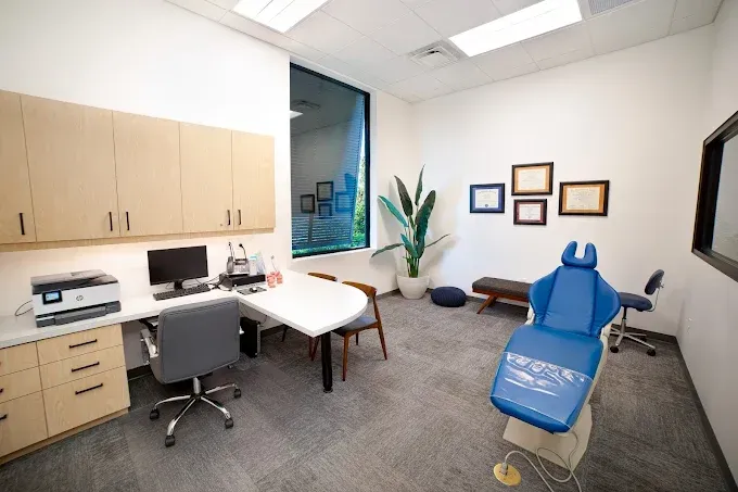 Doctor's office: blue dental chair, desk with computer, cabinets, window, potted plant, and framed art.