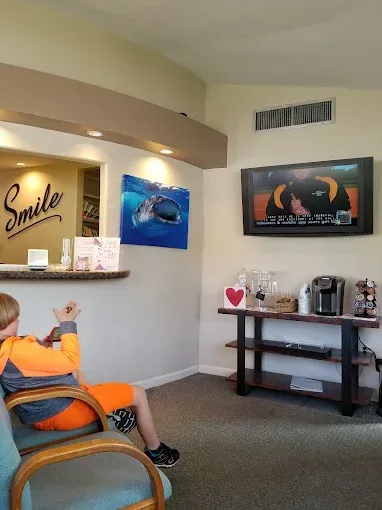 Boy in orange clothing waits in a dental office. TV playing, coffee station, reception desk.