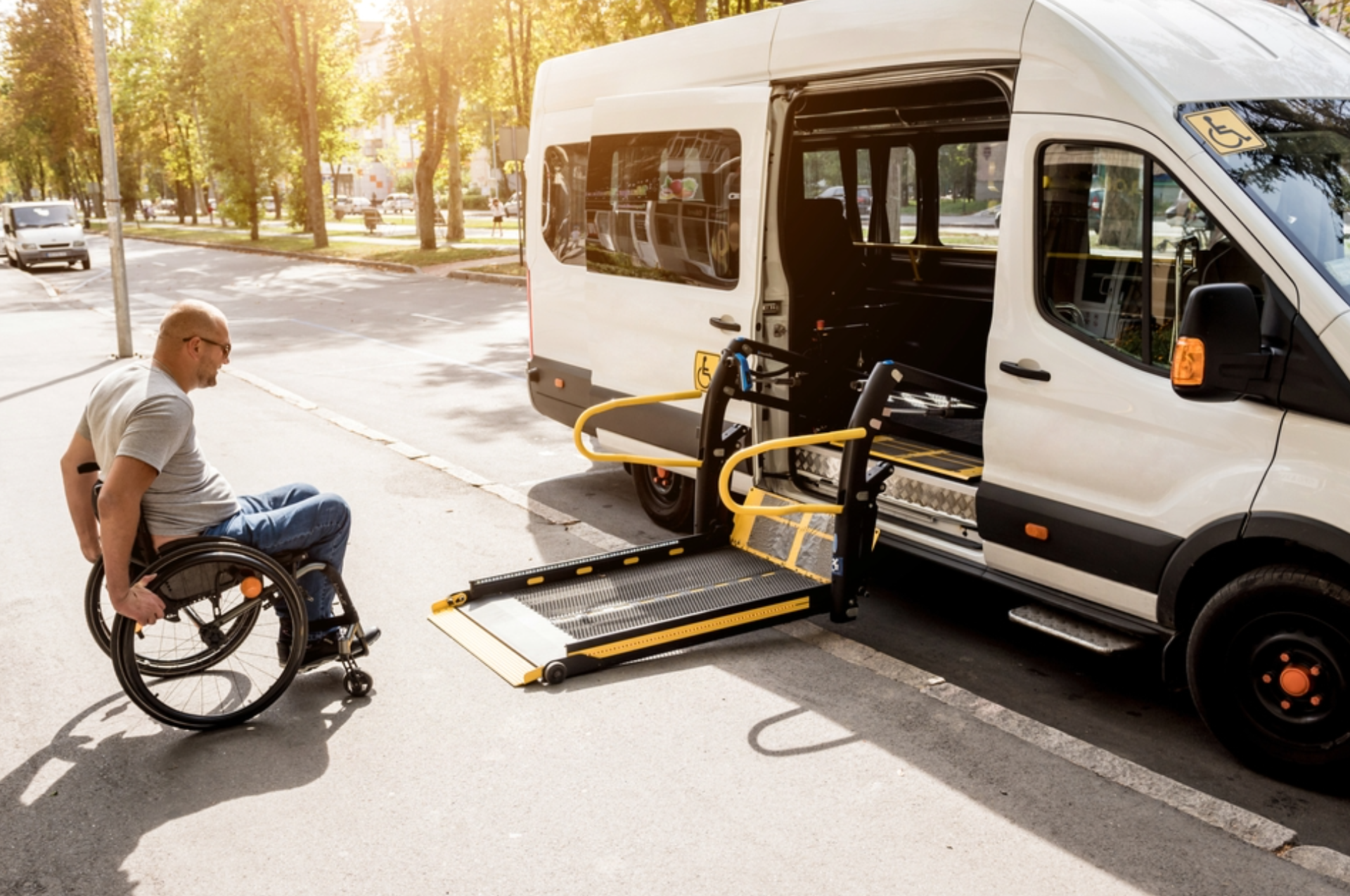 Man in wheelchair approaching a white accessible van with a deployed ramp on a sunny street.