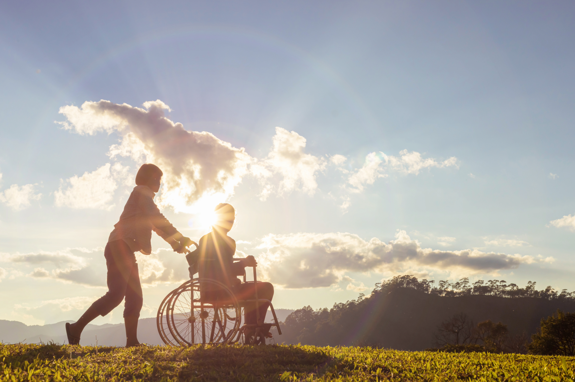 Woman pushing a person in a wheelchair through a sunny field; sunlight, hills, and clouds in the background.