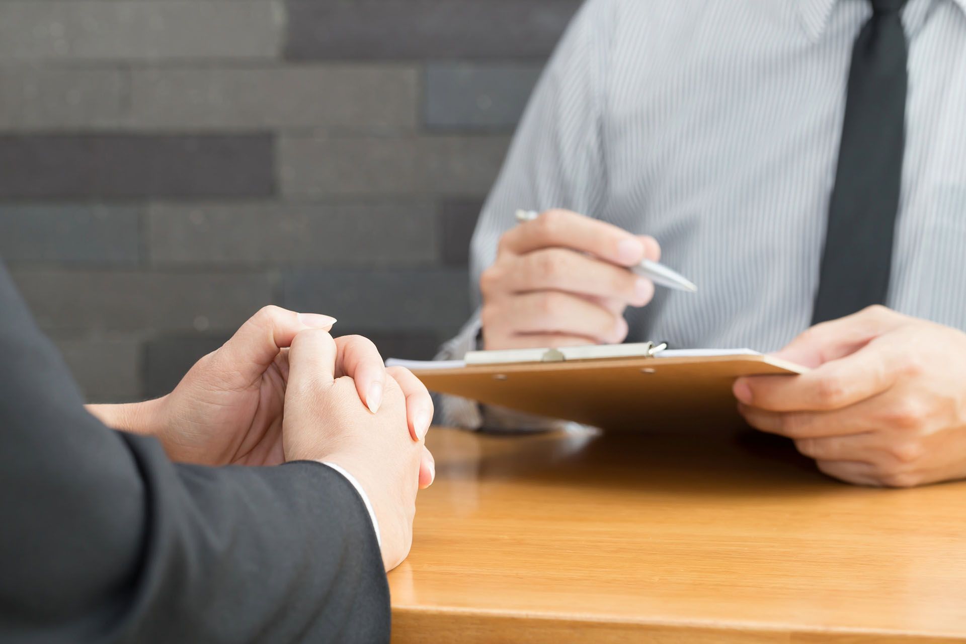 Person in suit at a desk, hands clasped, facing another person with a clipboard.