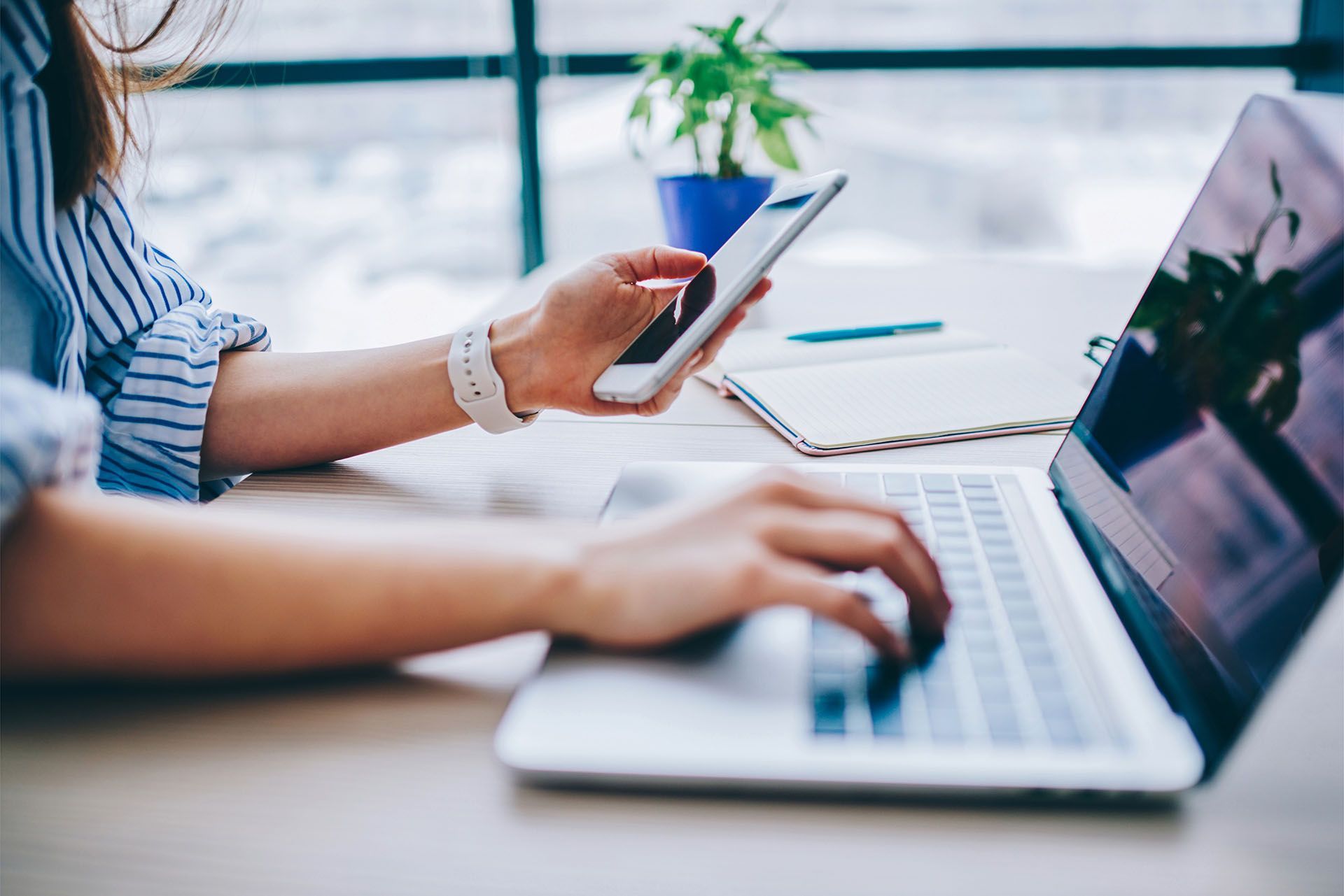 Woman using a laptop and smartphone at a desk near a window with a plant.