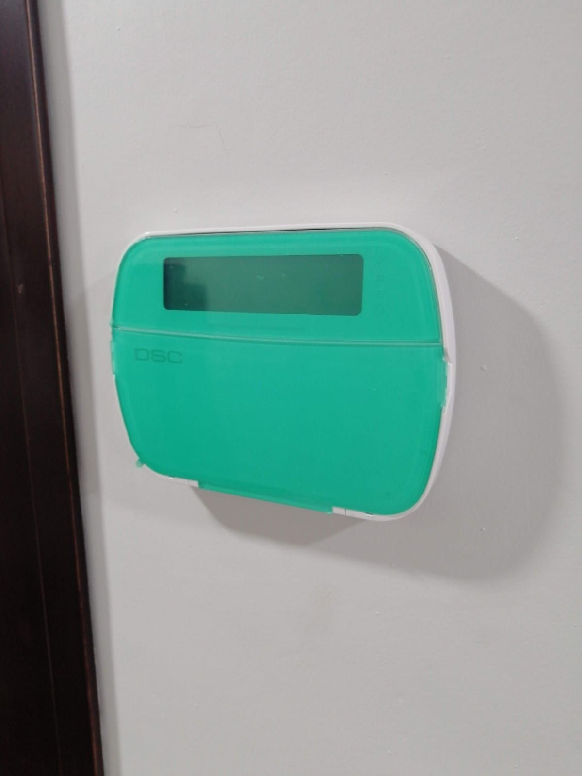 Una caja rectangular de color verde azulado para montar en la pared, con una ventana de visualización transparente, montada sobre una pared blanca.