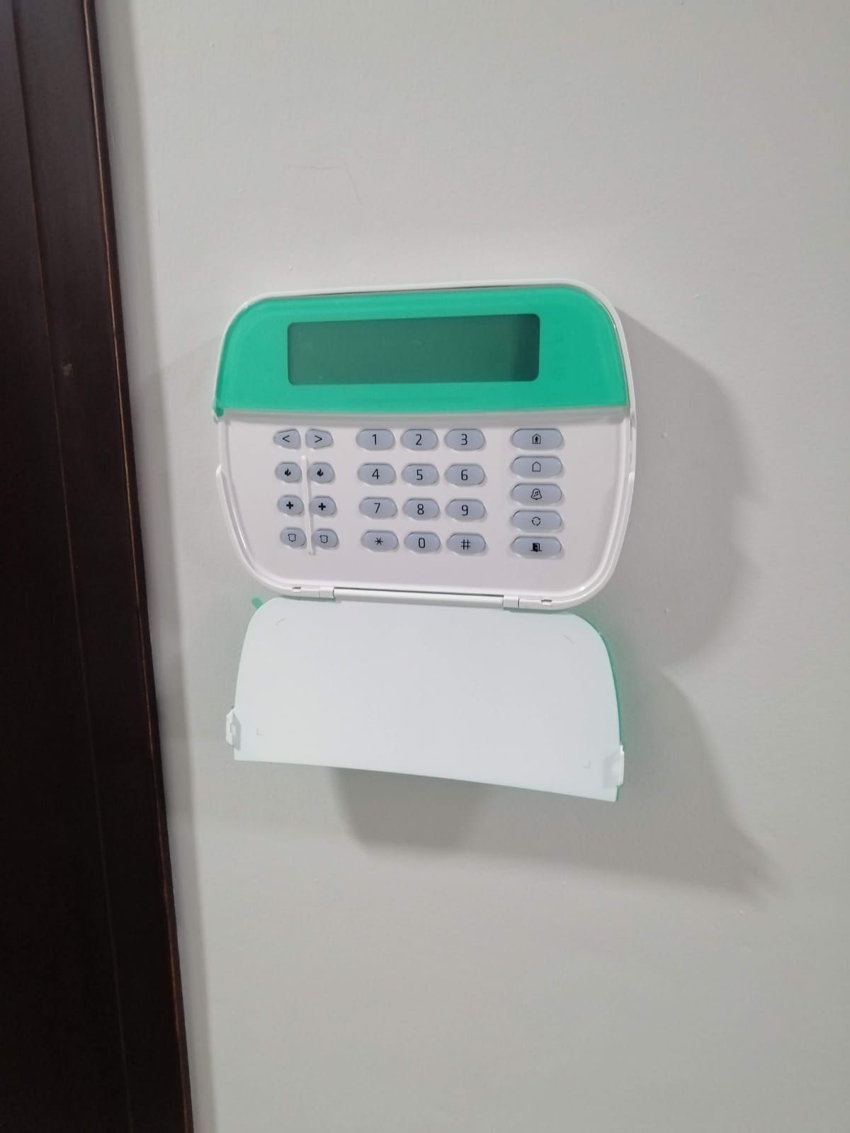 Teclado de alarma de seguridad sobre una pared blanca, con teclado numérico y pantalla, con detalles en color verde azulado.
