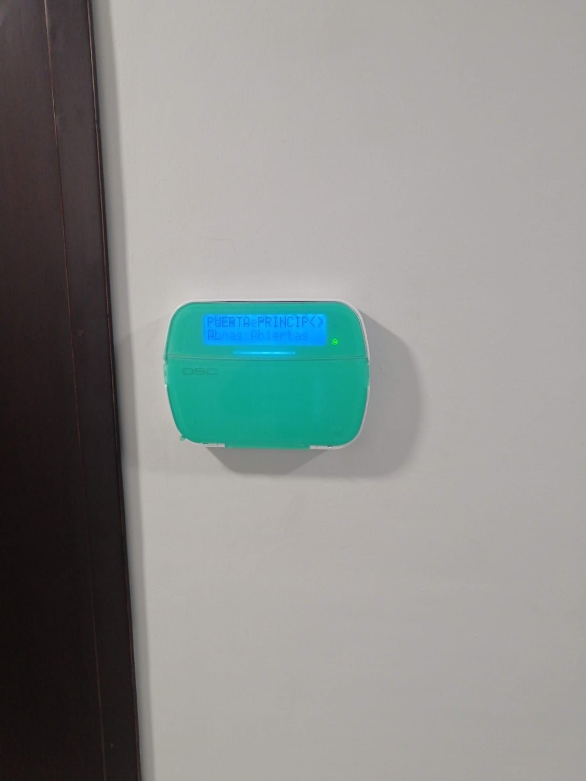 Dispositivo verde con pantalla azul sobre una pared blanca junto a una puerta marrón.