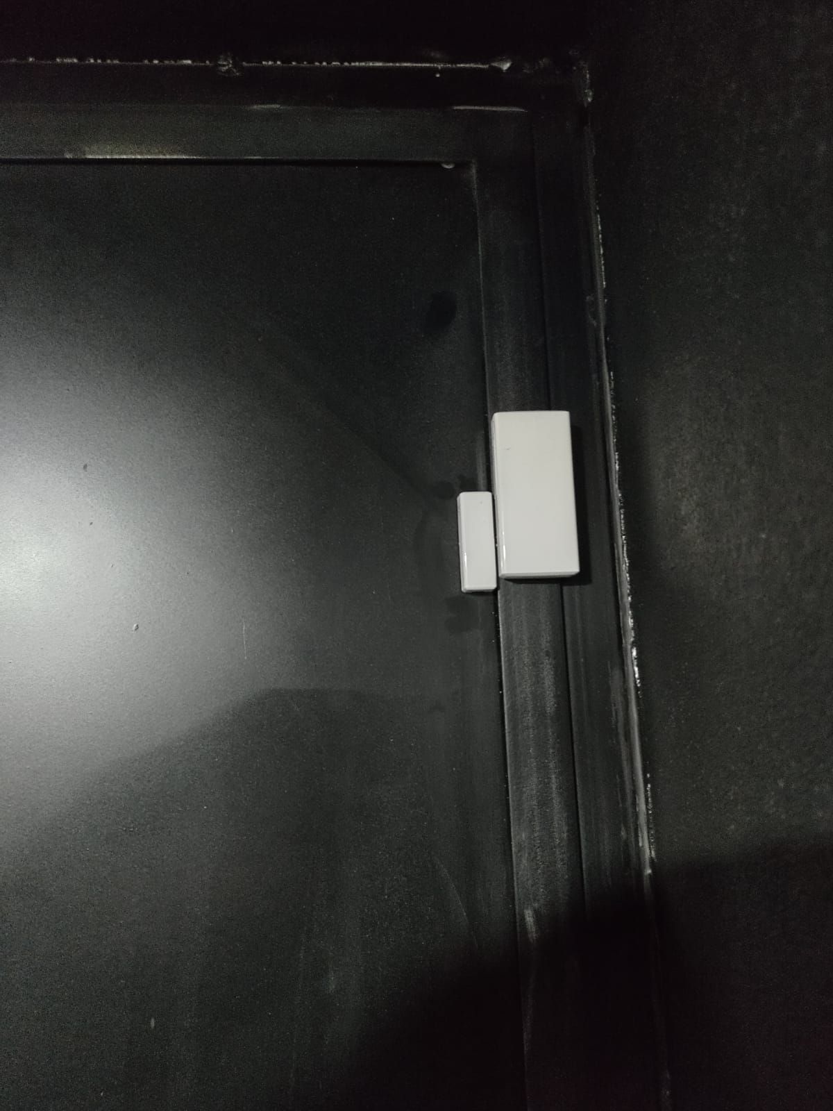 Sensor de puerta magnético blanco sobre marco de puerta negro, que indica un sistema de seguridad.