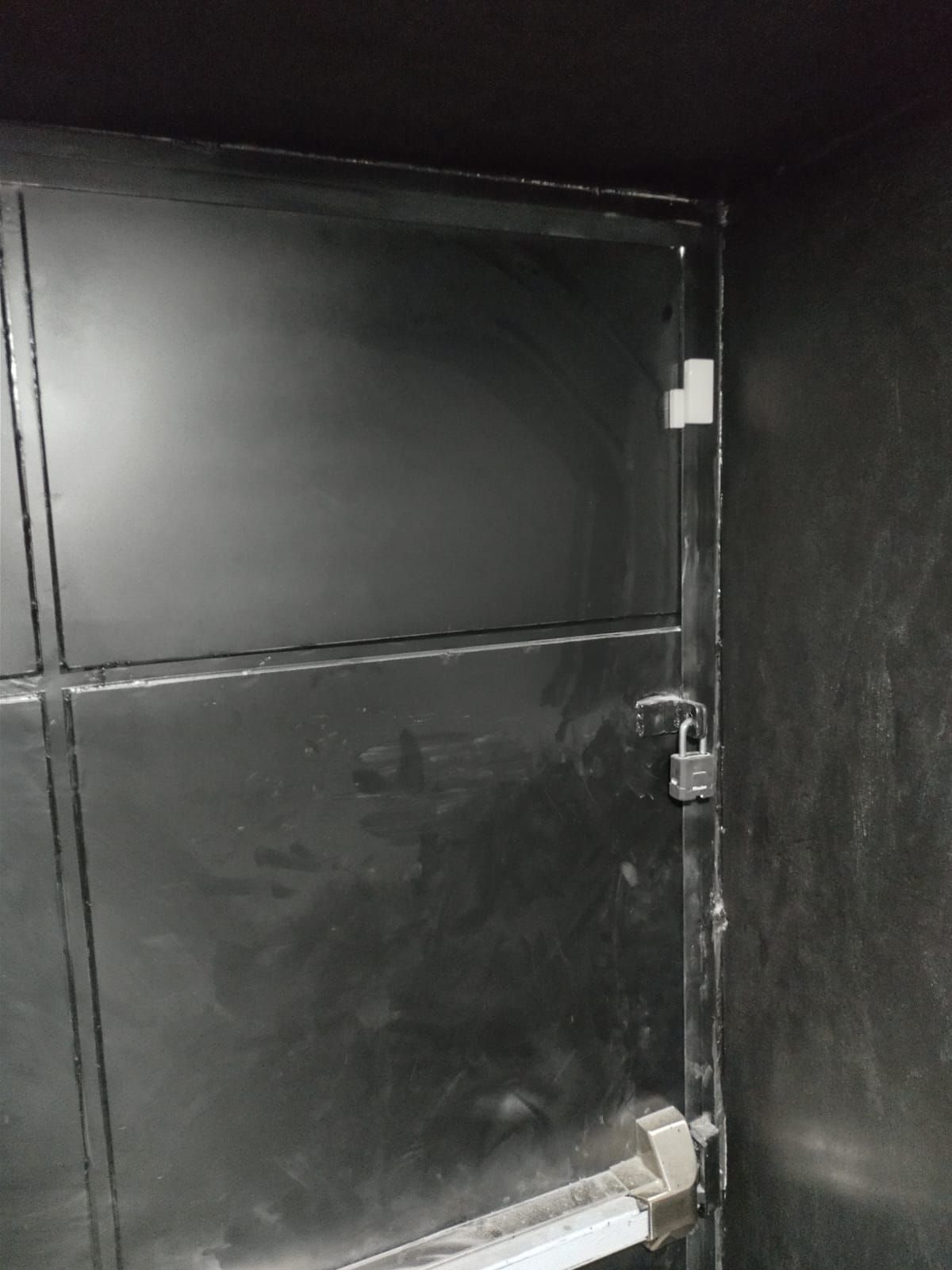 Puerta metálica de color negro con candado y barra de empuje en entorno oscuro.